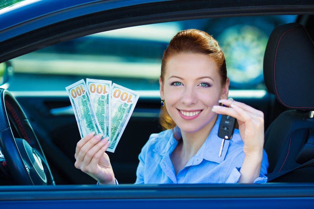 Image of a woman in the car smiling and holding the car key with bills of $100 on another hand after trade-in deal